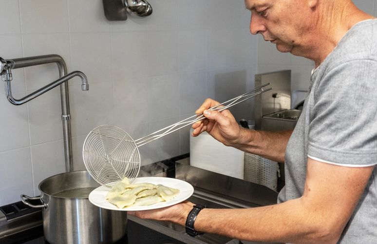 Ein Koch im grauen T-Shirt hält mit einer Schaumkelle frisch gekochte Maultaschen aus einem großen Topf und legt sie auf einen weißen Teller.