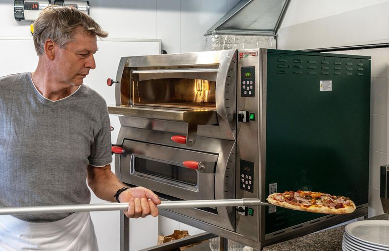 Ein Mann im grauen T-Shirt schiebt mit einer langen Metallschaufel eine frisch belegte Pizza in einen industriellen Pizzaofen.